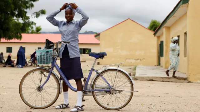 A girl stands astride her bike in front of a village compound