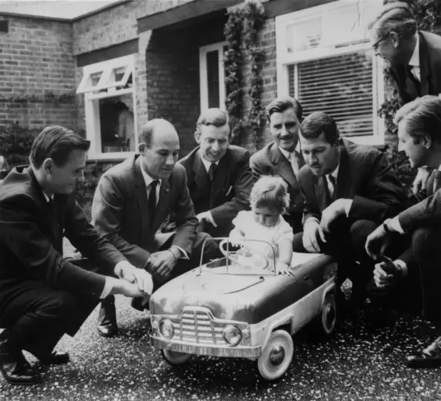 British motor racing driver Damon Hill sitting in a toy car at his christening watched by racing drivers (left to right): B McClaren, Stirling Moss, Tony Brooks, his father Graham Hill, Jo Bonnier and Wolfgang von Trips