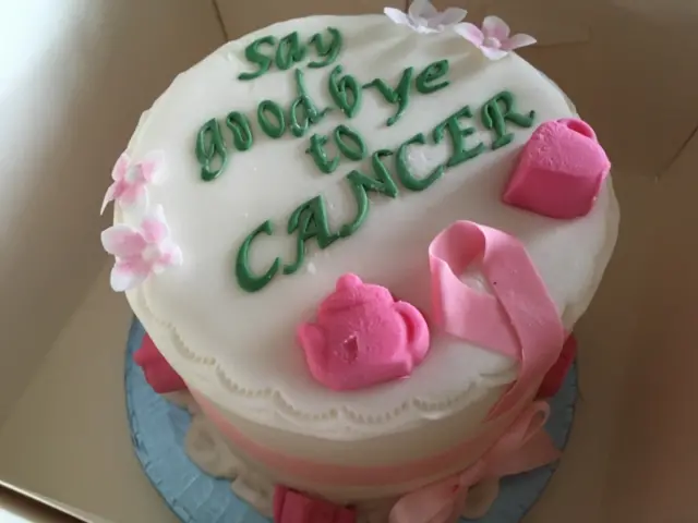 A white-iced cake, with pink iced decorations, and a green inscription saying Goodbye to Cancer