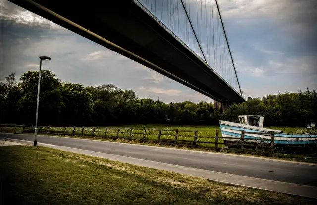 The Humber Bridge with a boat in a grassy field underneath.