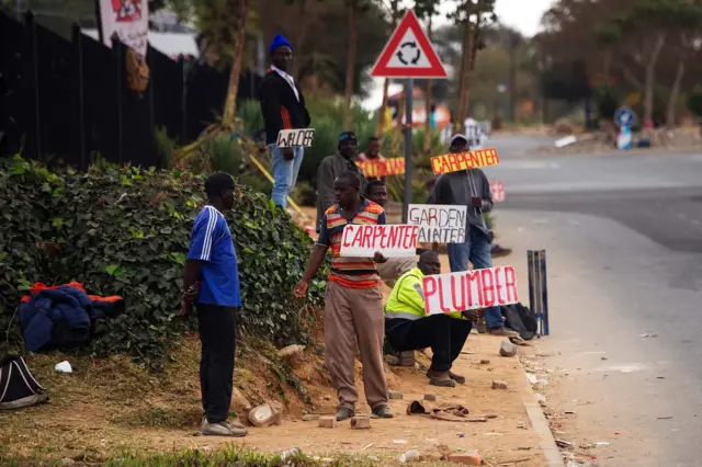 Job seekers wait on the side of a road holding placards reading their specialisation