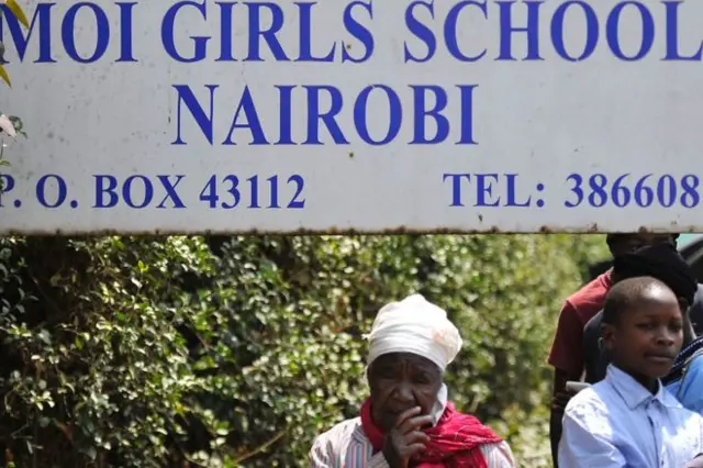 An elderly woman stands outside the school next to a sign bearing its name