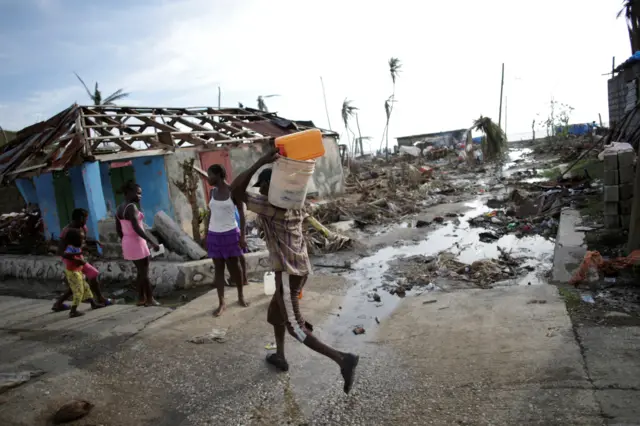 man carries containers on back past house with no roof; new wasteland in background