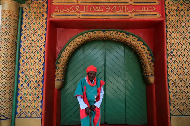 An elderly guard stands in front of an ornate doorway