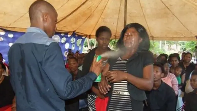 A man spraying insecticide in the face of a woman before a congregation.