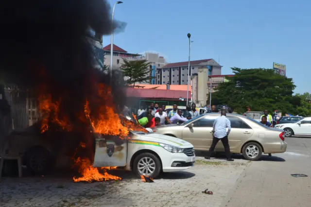 People watch a police vehicle as it goes up in flames following clashes with supporters of Islamic Movement of Nigeria (IMN) protesting against the imprisonment of their leader Ibraheem Zakzaky,