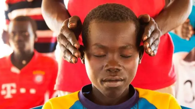 A boy sits with his eyes closed as another person holds his hands close to his head in a trauma healing session.