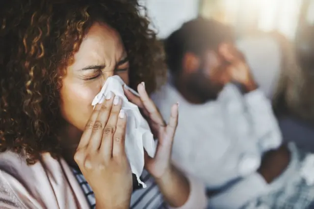 Woman blowing her nose with her boyfriend in the background