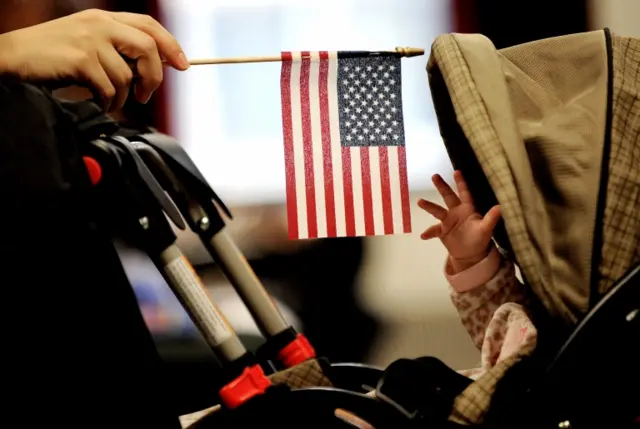 A baby reaches for an American flag held by her mother during naturalization ceremony