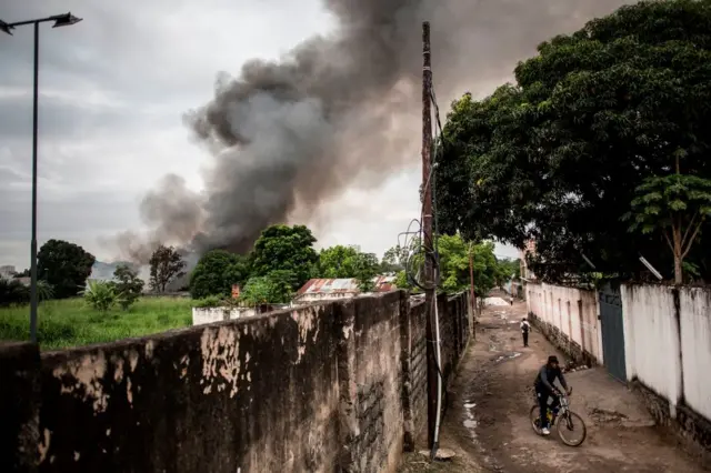 A man rides his bike as smoke rises from a fire at the independent national electoral commission's (CENI) warehouse on December 13, 2018 in Kinshasa, ten days ahead of presidential elections that have been foreshadowed by violence