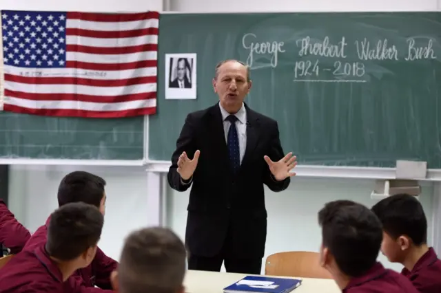 Teacher speaks to students with American flag and Bush photo in background