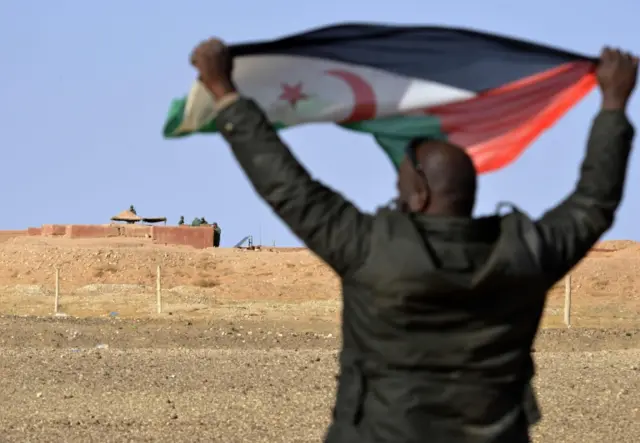 a Saharawi man holding up a Polisario Front flag in the Al-Mahbes area near Moroccan soldiers guarding the wall separating the Polisario controlled Western Sahara from Morocco.