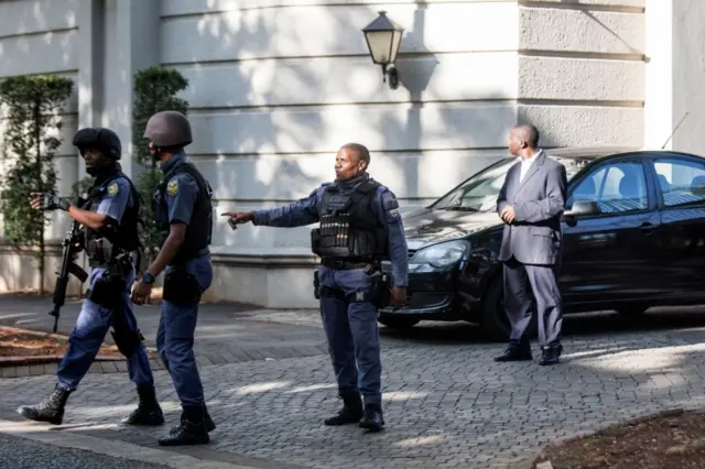 An armed South African Police member gestures as he leaves with colleagues the compound of the controversial business family Gupta while cars belonging to the Hawks, The Directorate for Priority Crime Investigation, are stationed outside, in Johannesburg, on February 14, 2018.