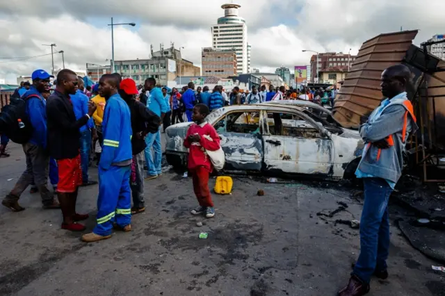 A crowd gathers around a burnt out shell of a car torched outside Harare Central Police Station on February 23, 2018, in Harare