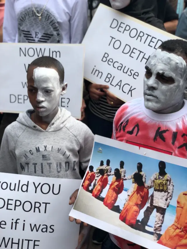 A close shot of two protesters wearing white facepaint and holding placards - one reads 'would you deport me if I was white'?