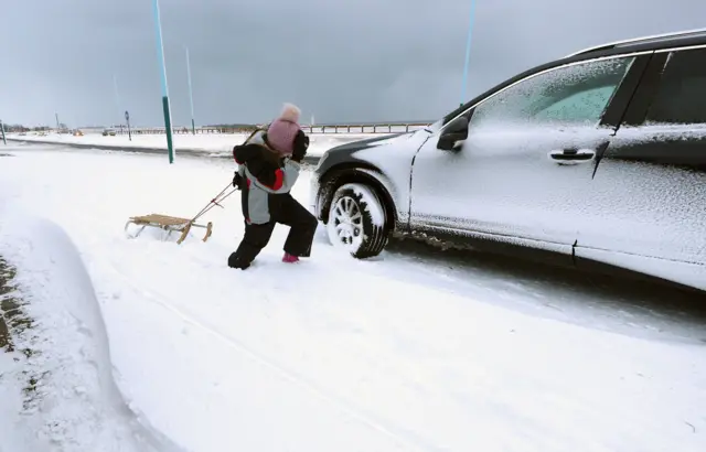 A youngster pulls a sled along a street alongside a snow-covered parked car