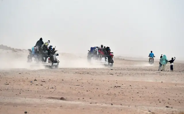 A woman and children wave to migrants sitting on pick-up trucks, holding wooden sticks tied to the vehicle to avoid falling from it, as they leave the outskirts of Agadez for Libya