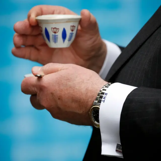 Rex Tillerson holds a cup of coffee in his hand, with his initials clearly visible embroidered onto his shirt cuff