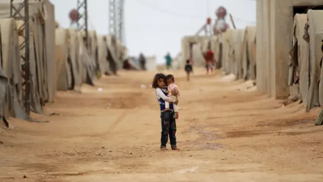 A girl carries a child inside a refugee camp in Syria