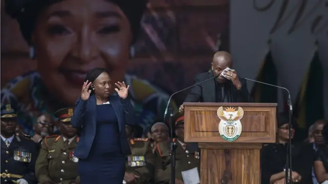 EFF leader Julius Malema stands a podium blotting his tears with a tissue while the sign language interpreter pauses to look at him with her hands aloft.