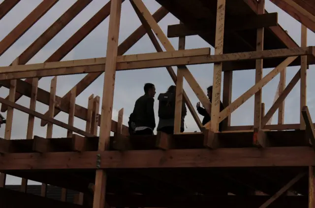 Three people at the top of a timber frame being made into a house.