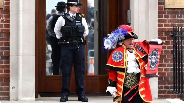 Town crier in traditional red regalia reads from a scroll and waves a bell