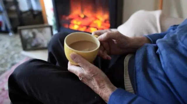 Elderly man sits in front of a fire with a cup of tea