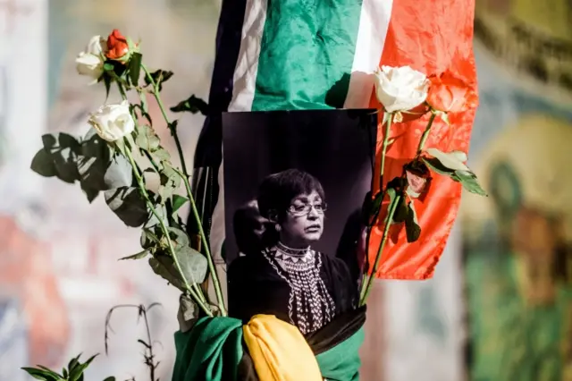 A black and white photograph of the late Winnie Madikizela-Mandela is surrounded by the South African and African National Congress (ANC) flags on a pole at the Old Durban Prison"s Human Rights wall