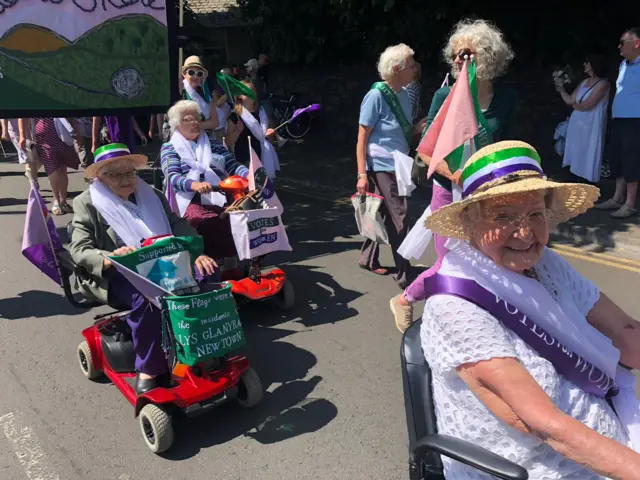 Women driving mobility scooters in the procession with their banners