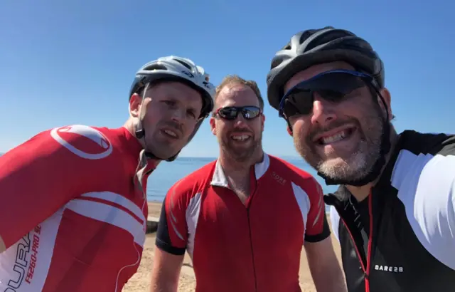 three men in cycling clothes on a beach