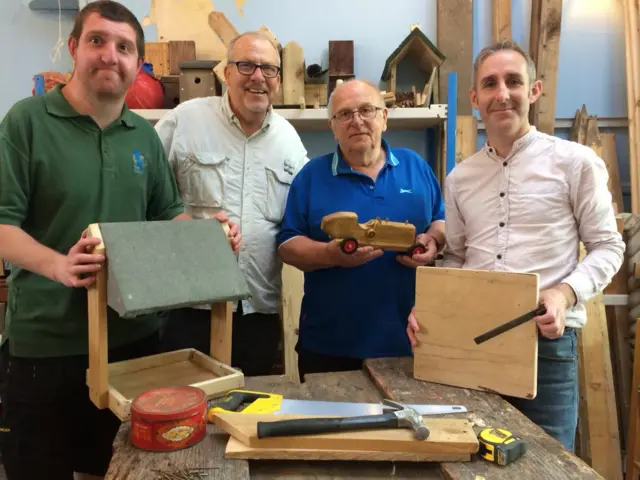 Four men stand with wooden creations by a work bench