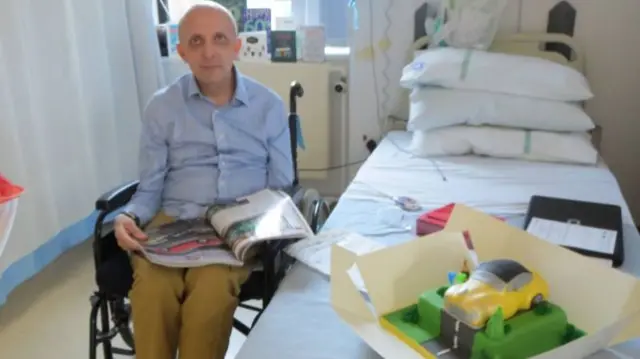 Dominic Excell sat in a hospital bay next to a bed with a cake on it.