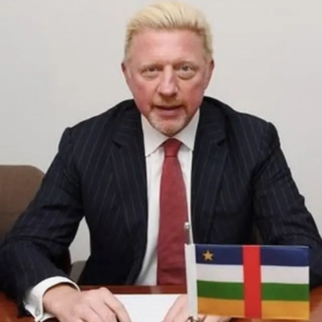 Boris Becker sits at a desk with the CAR national flag