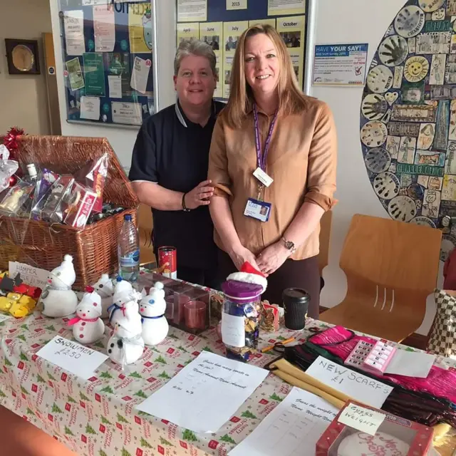 Two people stood behind a table which has cakes, scarfs and cuddly toys available to buy