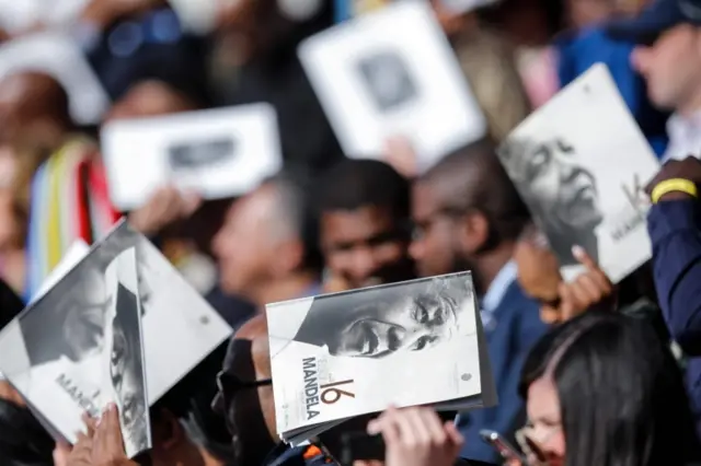 People use a brochure with the image of former South African leader Nelson Mandela to shade their heads from the sun during the Annual Nelson Mandela Lecture which will be given by the former US president at the Wanderers Cricket Stadium in Johannesburg on July 17, 2018.