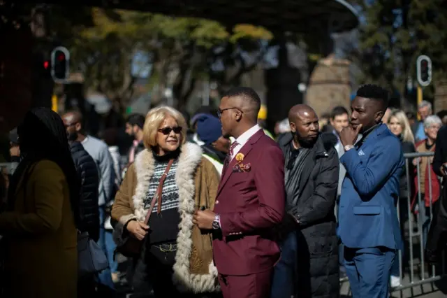 A man dressed in a maroon suit chats with a woman in the queue