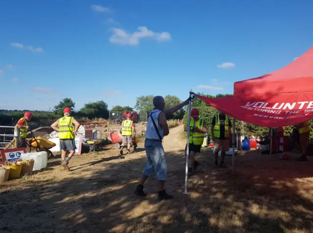 People in High Vis waistcoats and red helmets putting up a gazebo and pushing wheel barrows