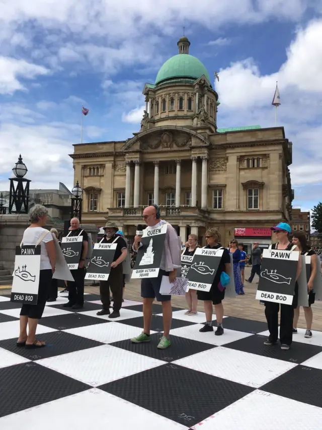 People stand on a chess board with A-boards describing the chess piece the represent in front of Hull City Hall