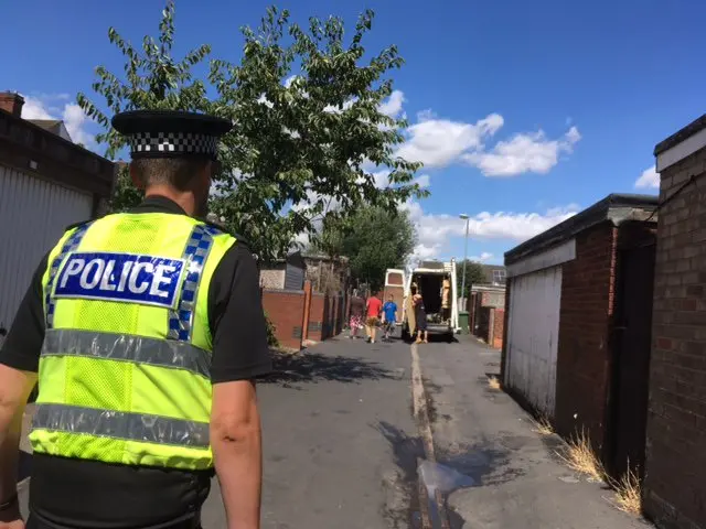 A police officer walks along road towards van in the distance with open doors and a number of people nearby