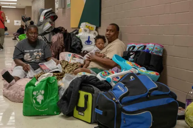 Family sits among their belongings on the floor of a shelter