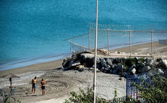 A picture taken on September 4, 2018 shows men gesturing towards a section of the border fence encircling Spain"s North African enclave of Ceuta which lies on the Strait of Gibraltar, surrounded by Morocco.