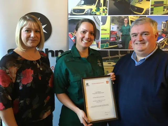 three people, the woman in the middle is holding a certificate