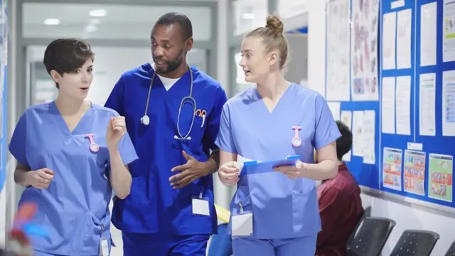 Three hospital staff in blue scrubs walk down a corridor talking