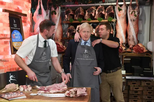 Prime Minister Boris Johnson (centre) has help putting on an apron during a visit to Dart Farm Village in Topsham, Exeter, while on the General Election campaign trail.