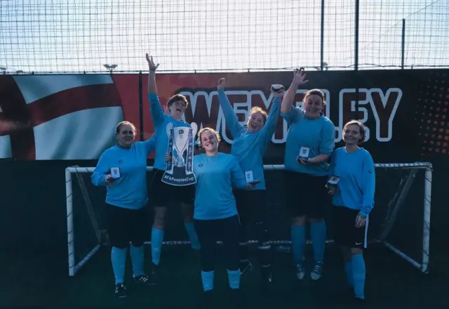 A FA People's Cup Female Walking football team celebrate with a cardboard cut out Trophy because they have made it to the next round