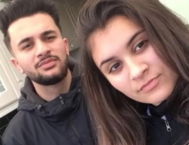 a young woman standing in front of her brother, both looking to camera