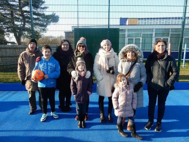 A group of family and friends of different ages smiling to camera standing on a 5-a-side pitch