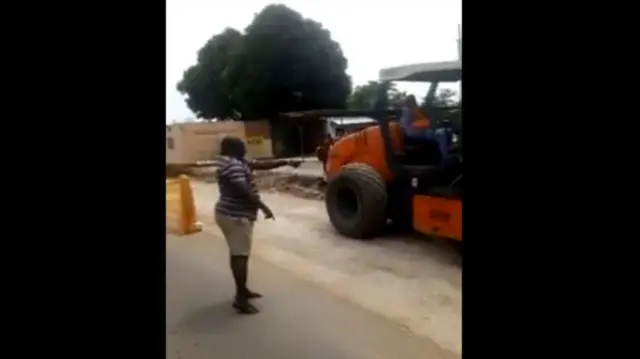 The minister is seen standing on the road and pointing at a man operating a vehicle