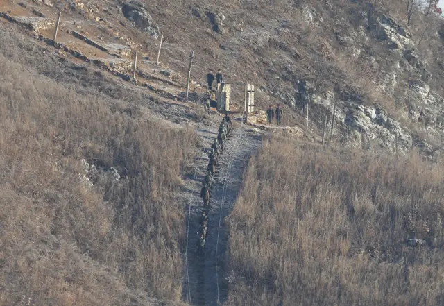 South Korean soldiers, wearing yellow head bands, and North Korean army soldiers move to inspect the dismantled North Korean guard post inside the Demilitarized Zone (DMZ) in the central section of the inter-Korean border as South Korean army soldiers watch in Cheorwon, Korea, Dec. 12, 2018.