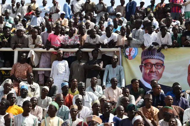 Supporters sit beside a banner of candidate of the ruling All Progressives Congress (APC) President Mohammadu Buhari during his presidential campaign rally at the Sanni Abacha Stadium in Kano, on January 31, 2019.